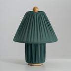 Dark green Clérachic pleated lamp, unlit, with a wooden ball finial and a wooden foot, on a pale grey surface.
