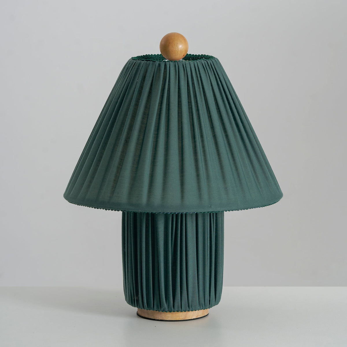 Dark green Clérachic pleated lamp, unlit, with a wooden ball finial and a wooden foot, on a pale grey surface.