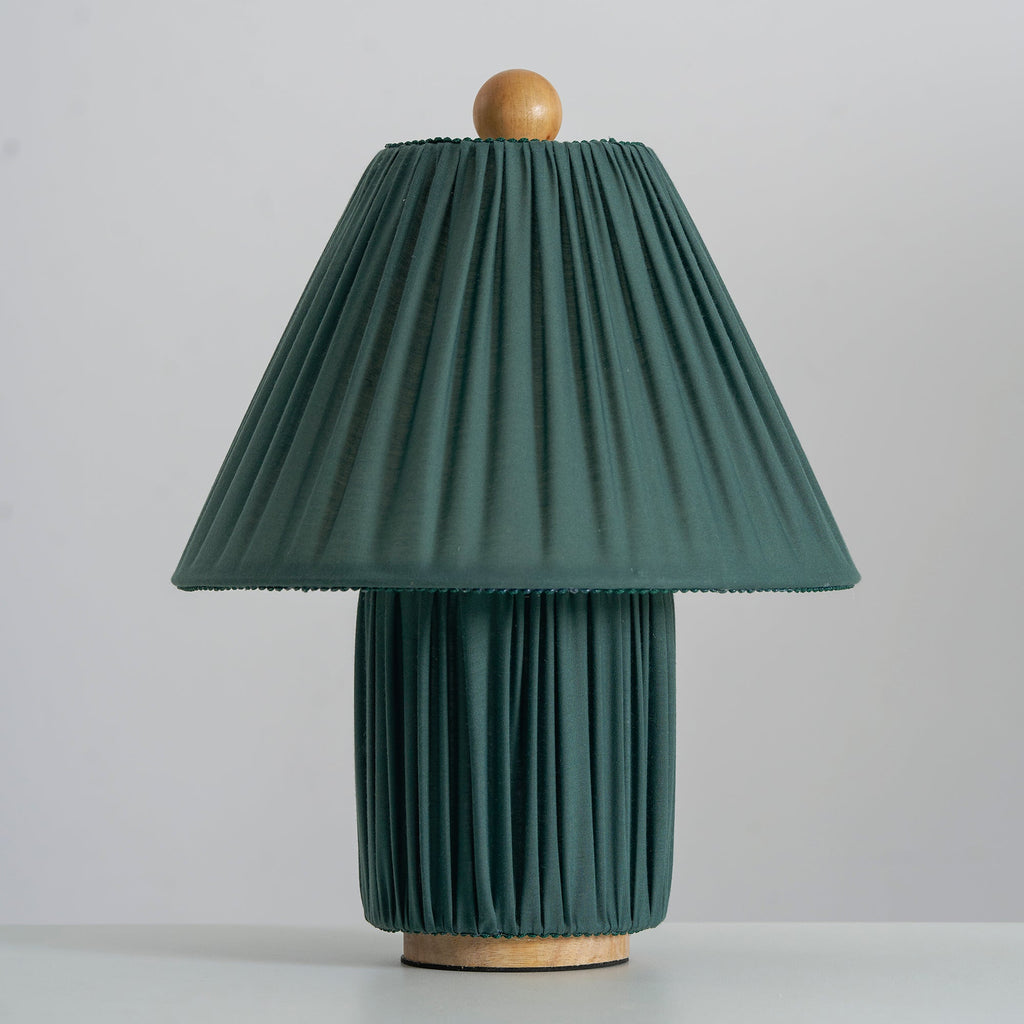 Unlit dark green Clérachic table lamp seen head-on, its pleated shade edged with a fine trim, on a pale grey surface.