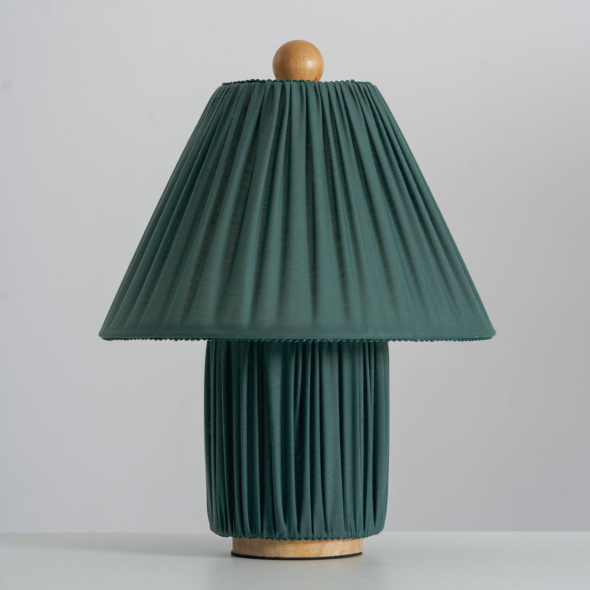 Unlit dark green Clérachic table lamp seen head-on, its pleated shade edged with a fine trim, on a pale grey surface.