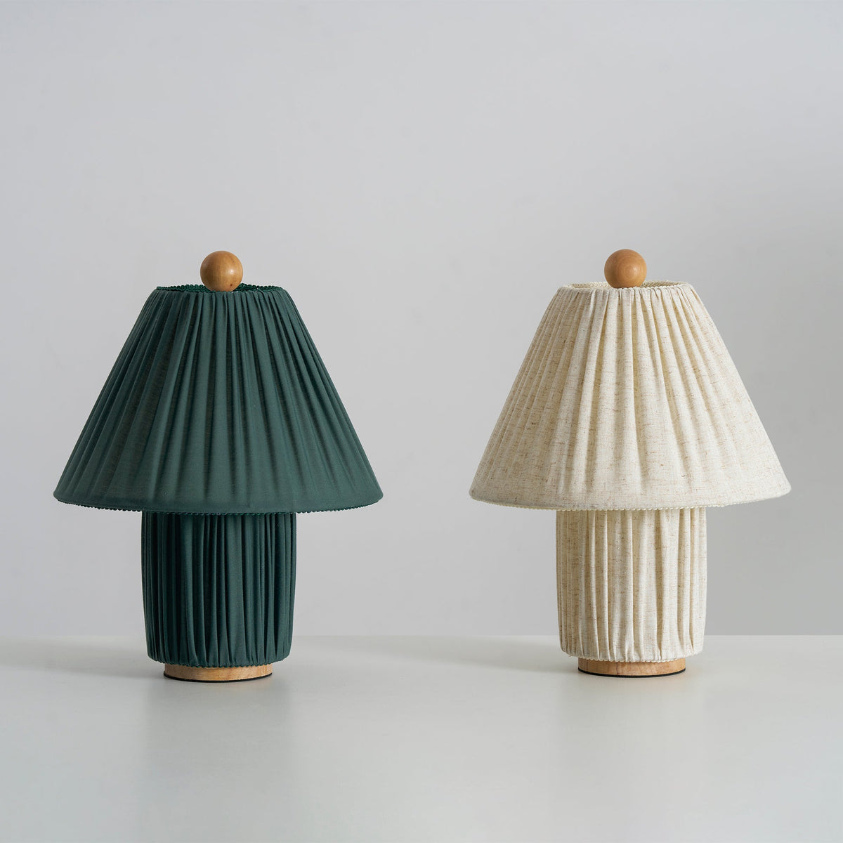 Two Clérachic pleated lamps side by side, one dark green and one linen, both unlit, on a pale surface.