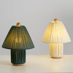 The dark green and linen Clérachic pleated lamps side by side, both lit, the linen shade glowing a warm yellow.