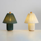 From farther back, a dark green and a linen Clérachic pleated lamp stand together, both lit, on a pale grey surface.
