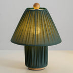 Dark green Clérachic pleated lamp, lit, its shade showing the pleats and a soft glow, on a pale surface.