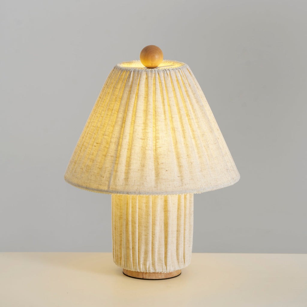 Linen Clérachic pleated lamp lit warm, its shade glowing and its pleated base lit from within, on a pale surface.