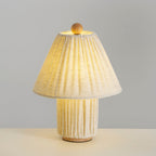 Linen Clérachic pleated lamp lit warm, its shade glowing and its pleated base lit from within, on a pale surface.