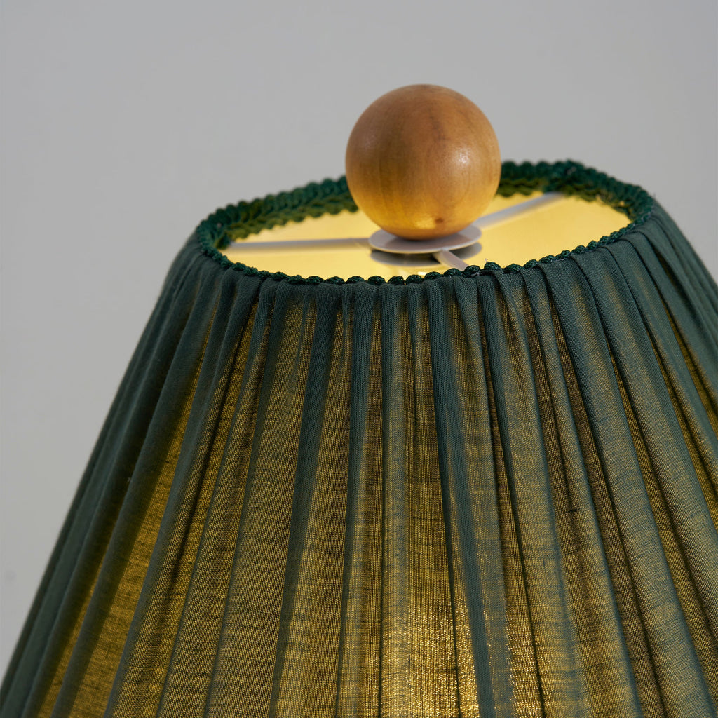 Close view of the lit top of a dark green Clérachic lamp shade, with a wooden ball finial and a trimmed edge.