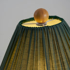 Close view of the lit top of a dark green Clérachic lamp shade, with a wooden ball finial and a trimmed edge.