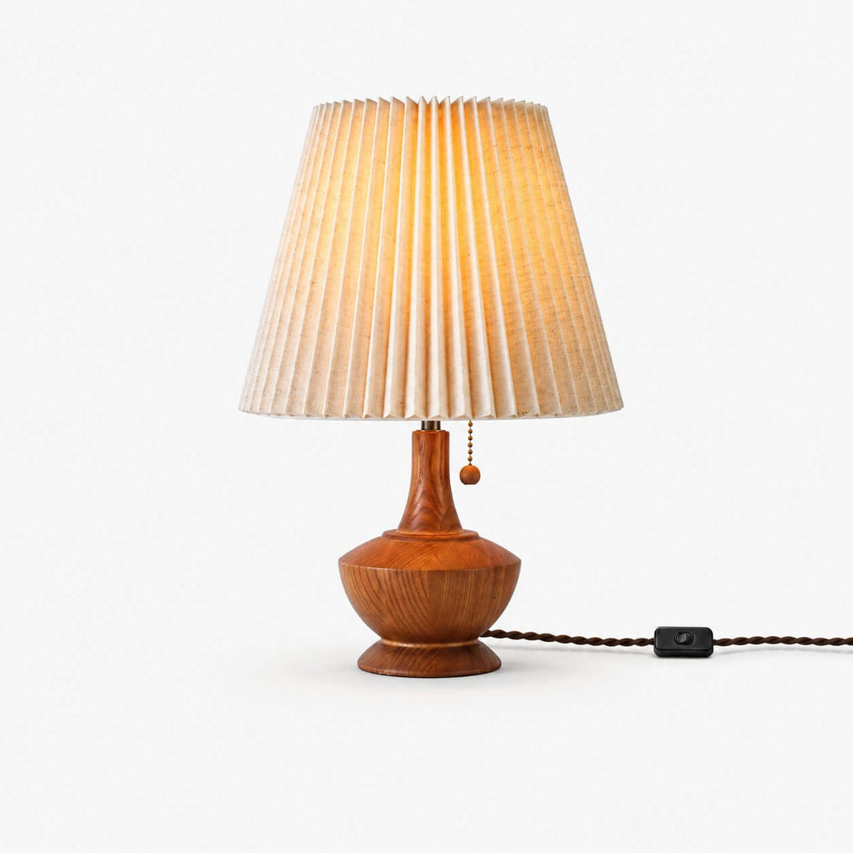This Clérachic table lamp on a white backdrop, a pleated beige fabric shade over a rounded wood base with a pull chain and a braided cord with an inline switch.