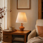 Lit Clérachic pleated table lamp on a wooden side table beside a tray with a cup, with a cream armchair and linen curtains.