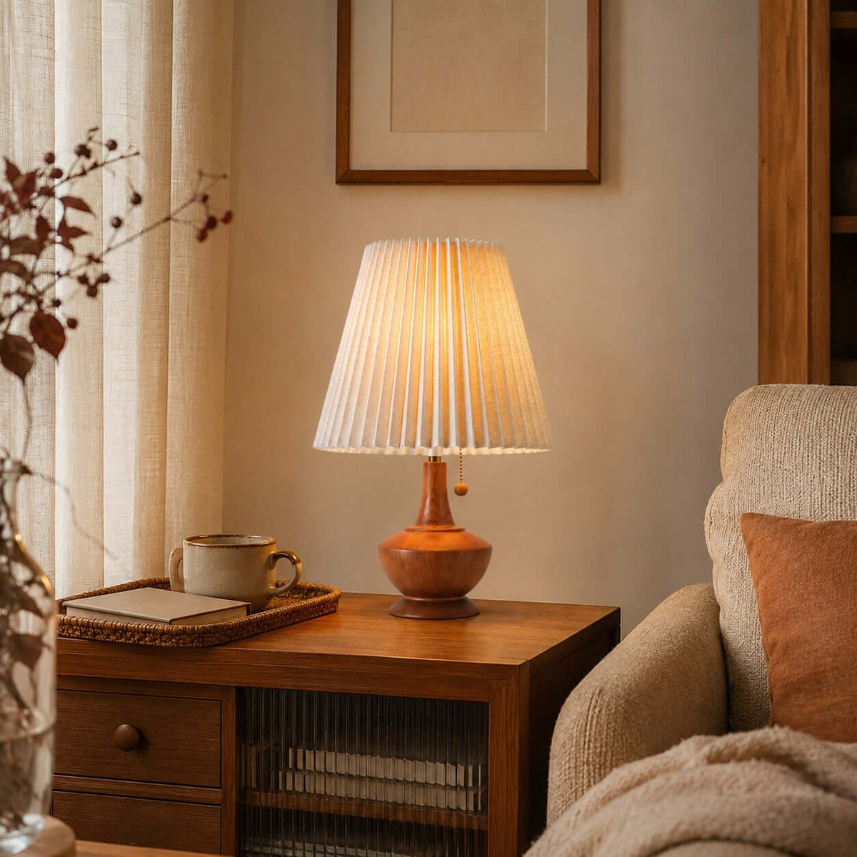Lit Clérachic pleated table lamp on a wooden side table beside a tray with a cup, with a cream armchair and linen curtains.