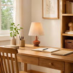 A Clérachic lamp on a light wood desk by a window, with an open notebook, a pen pot and a small plant in a vase.