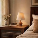 Clérachic lamp on a bedside cabinet with a ribbed glass door, next to a bed with linen bedding and a vase of pale roses.