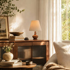 A Clérachic lamp on a wooden cabinet by a sunlit window, with a wooden bowl, a stack of books and a stone vase below.