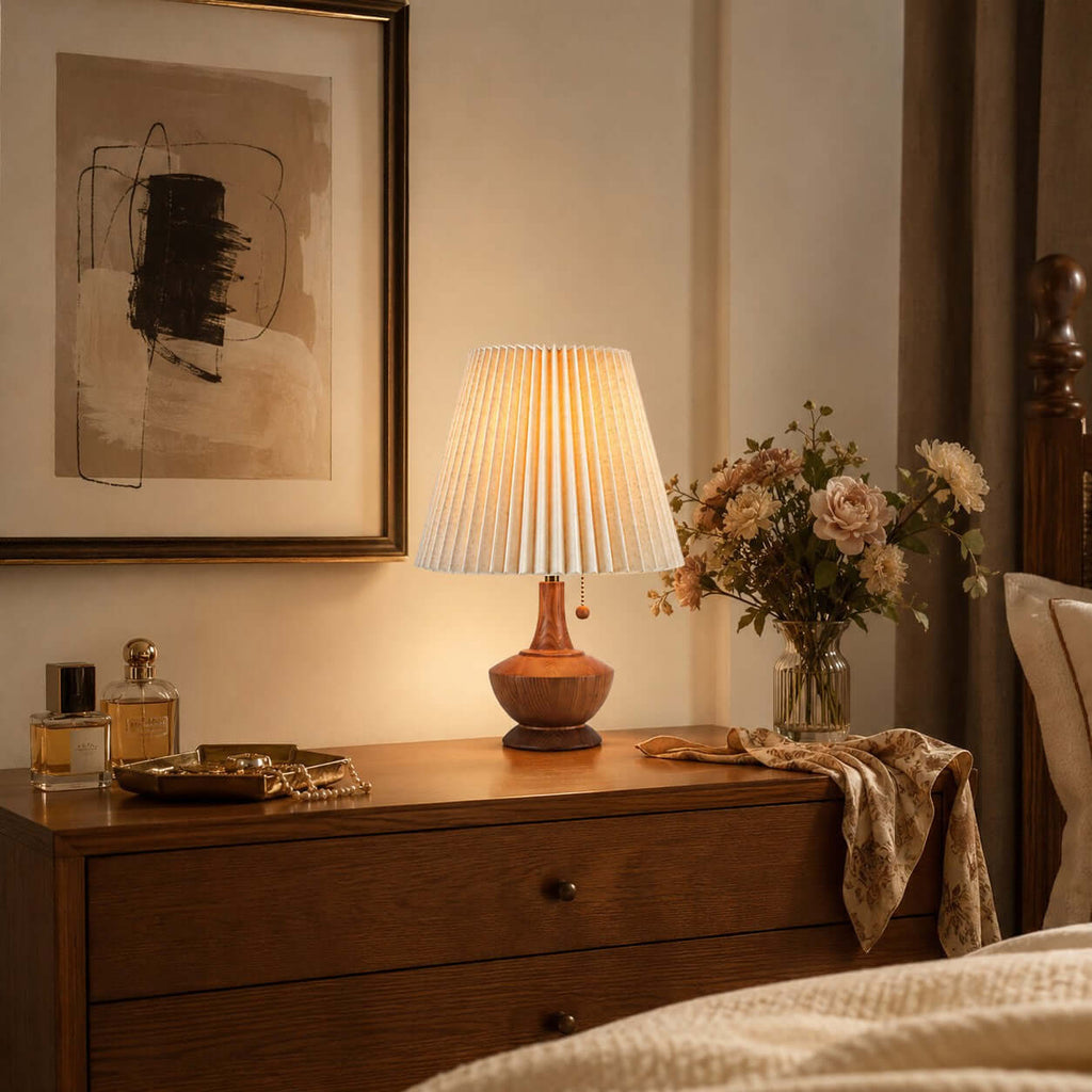 Clérachic pleated lamp on a chest of drawers below a large abstract print, beside perfume bottles and a vase of peach flowers.
