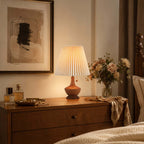 Clérachic pleated lamp on a chest of drawers below a large abstract print, beside perfume bottles and a vase of peach flowers.