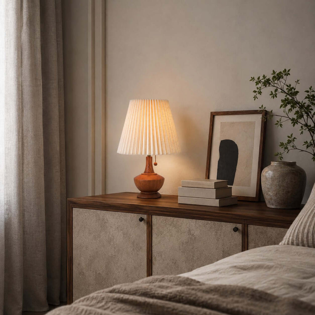 A Clérachic lamp on a low cabinet beside a framed print and a stone vase, with soft grey curtains and bed linen in the foreground.