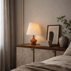 A Clérachic lamp on a low cabinet beside a framed print and a stone vase, with soft grey curtains and bed linen in the foreground.