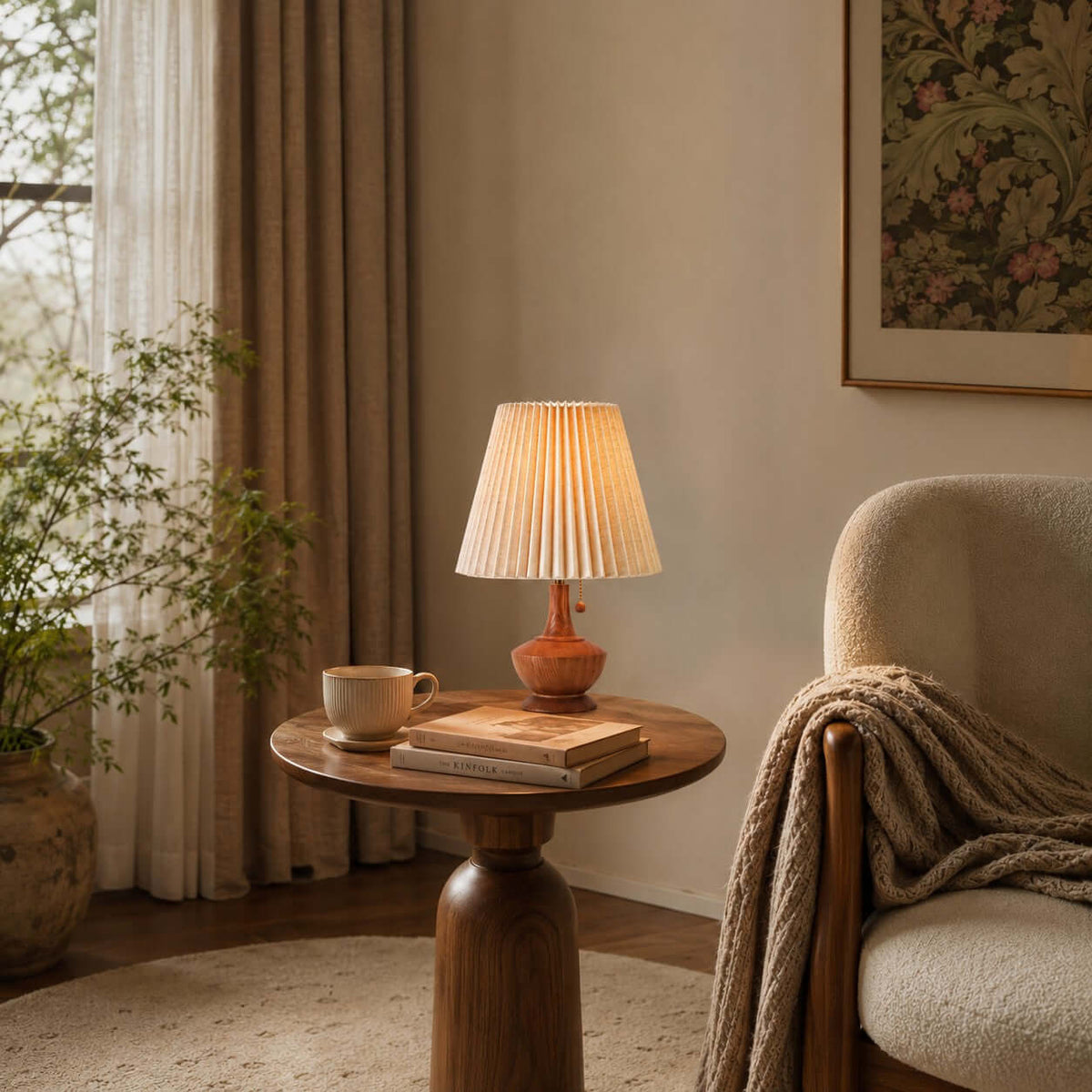 Clérachic table lamp on a round wooden side table with a cup and books, beside a bouclé armchair with a knitted throw.