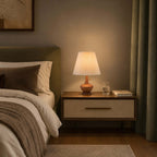 Clérachic lamp on a cream-fronted bedside table, with a glass of water and a book, beside a bed and olive-green curtains.