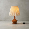 Close front view of the Clérachic table lamp on a plain grey floor and wall, showing the pleated shade, wood base and cord switch.