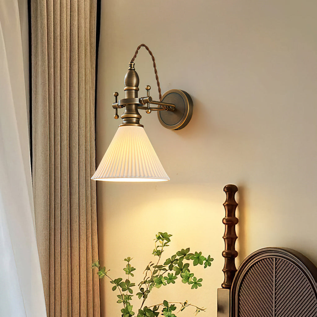 A Clérachic wall light with a pleated white cone shade glowing warmly beside linen curtains and a dark wooden bedhead.