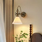 A Clérachic wall light with a pleated white cone shade glowing warmly beside linen curtains and a dark wooden bedhead.