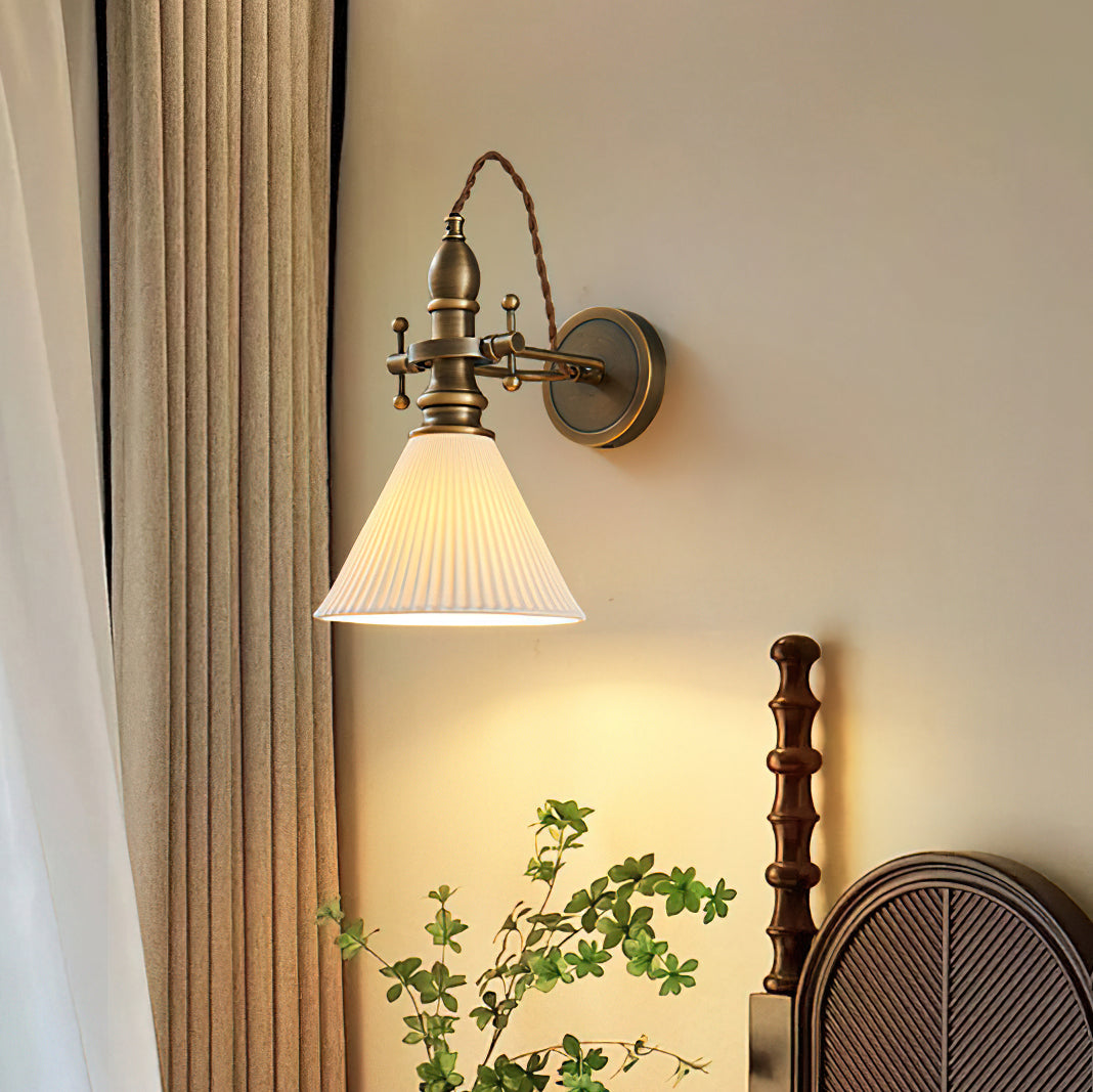A Clérachic wall light with a pleated white cone shade glowing warmly beside linen curtains and a dark wooden bedhead.