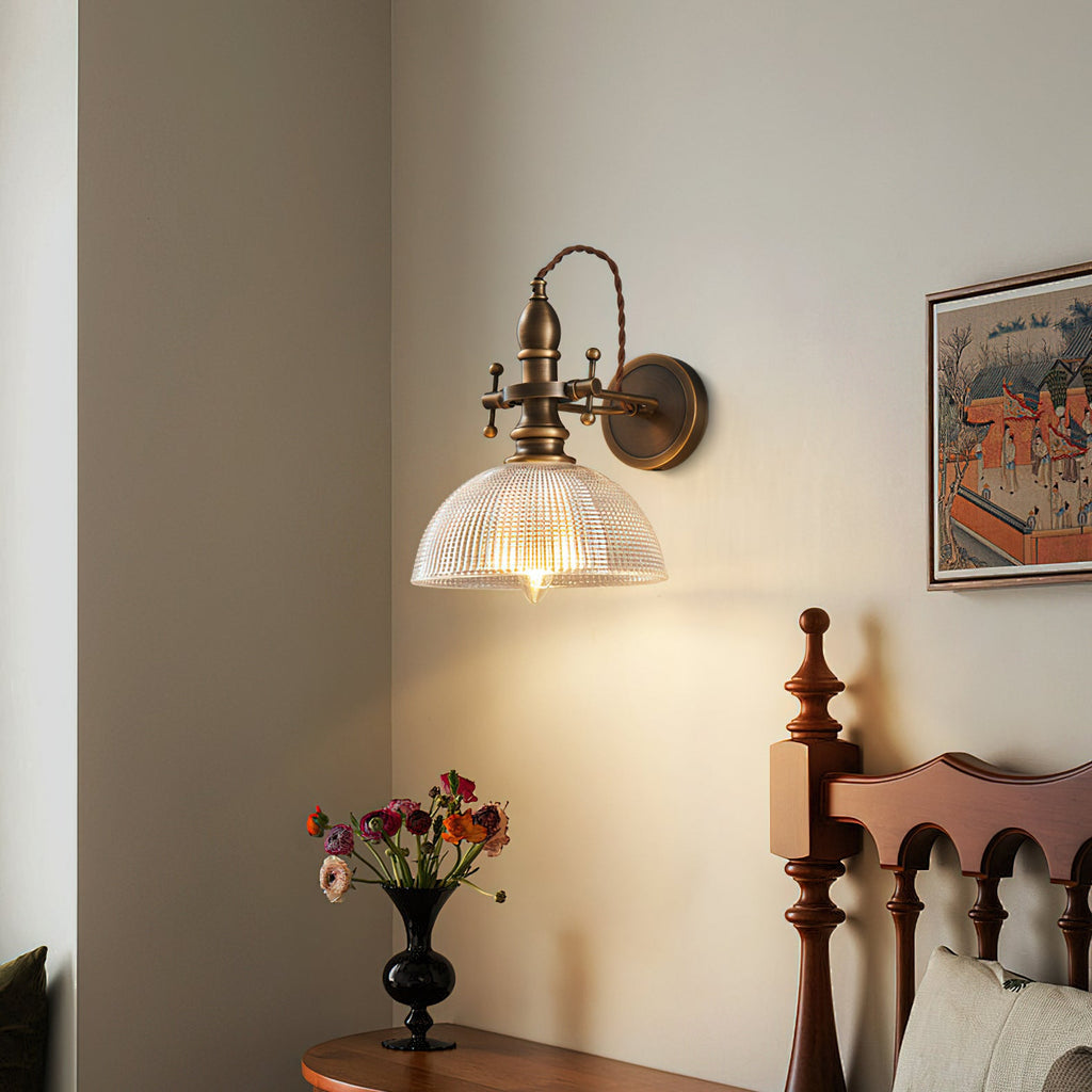 Clear ribbed-glass dome shade lit on a Clérachic brass wall light above a bedside table with a vase of flowers.