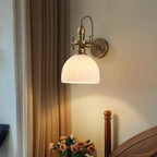 Ribbed white dome shade glowing on a Clérachic antique brass wall light, with roses on a table and curtains at the side.