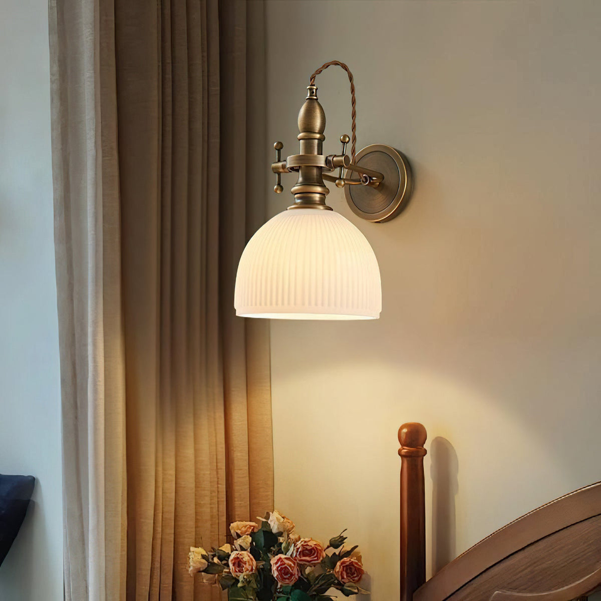 Ribbed white dome shade glowing on a Clérachic antique brass wall light, with roses on a table and curtains at the side.