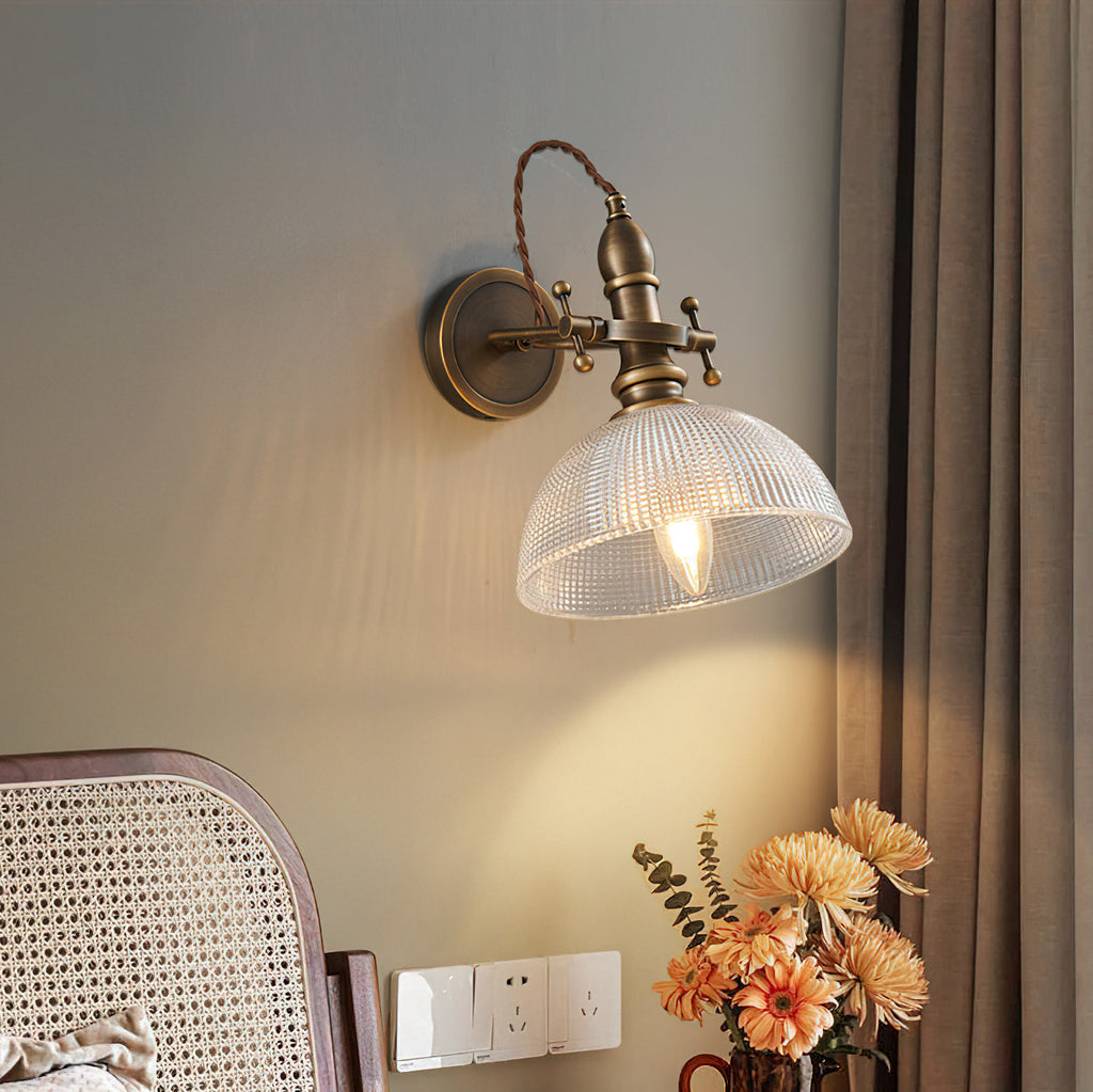 Clear textured-glass dome shade with a visible filament bulb on a Clérachic brass wall light, next to a rattan chair and orange flowers.