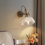 Clear textured-glass dome shade with a visible filament bulb on a Clérachic brass wall light, next to a rattan chair and orange flowers.