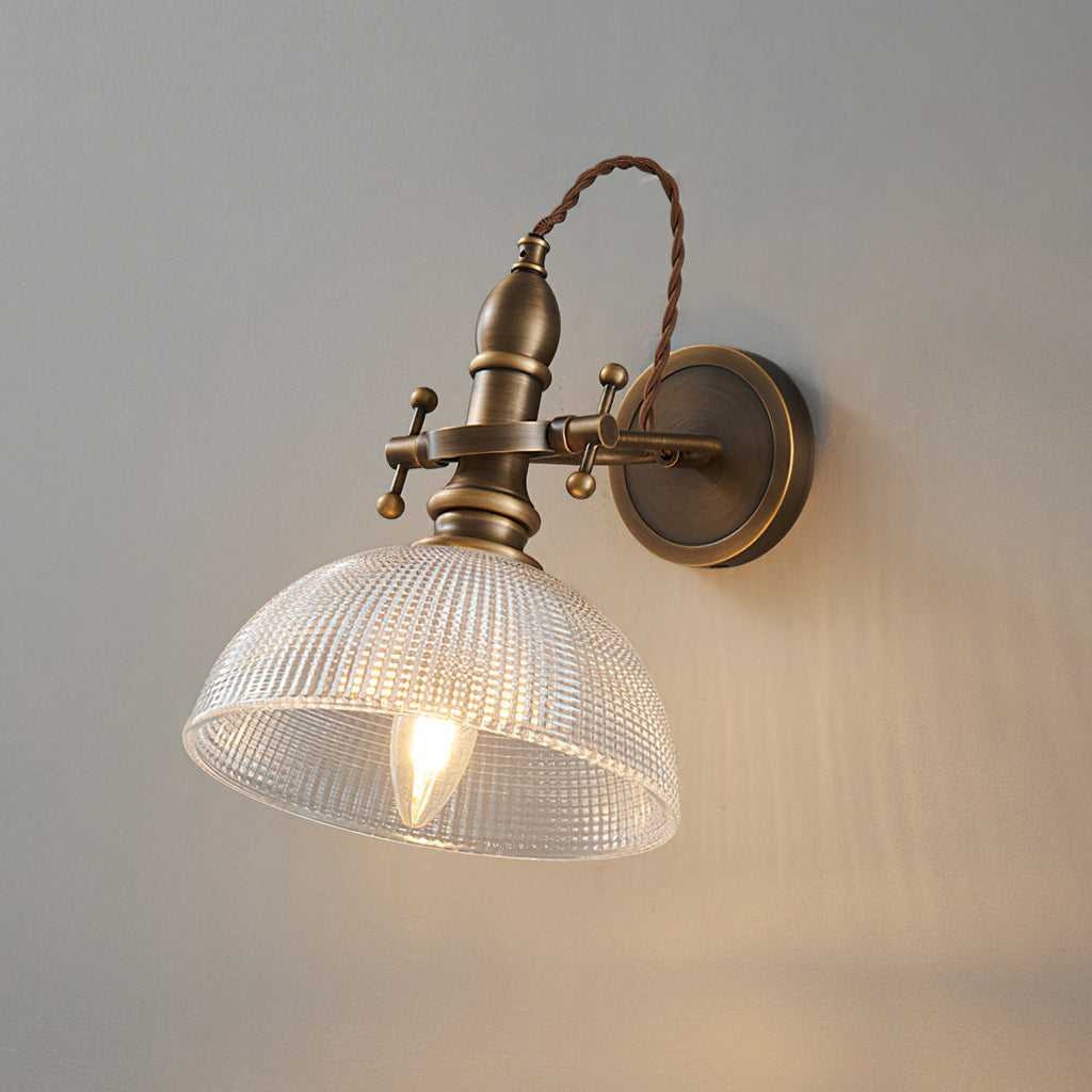 Close view of a ribbed clear-glass dome and lit filament bulb on this Clérachic brass wall light, against a grey wall.