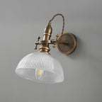 Unlit clear ribbed-glass dome shade showing the filament bulb inside, on a Clérachic antique brass wall light against grey.