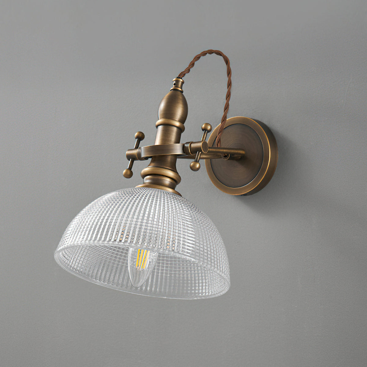 Unlit clear ribbed-glass dome shade showing the filament bulb inside, on a Clérachic antique brass wall light against grey.