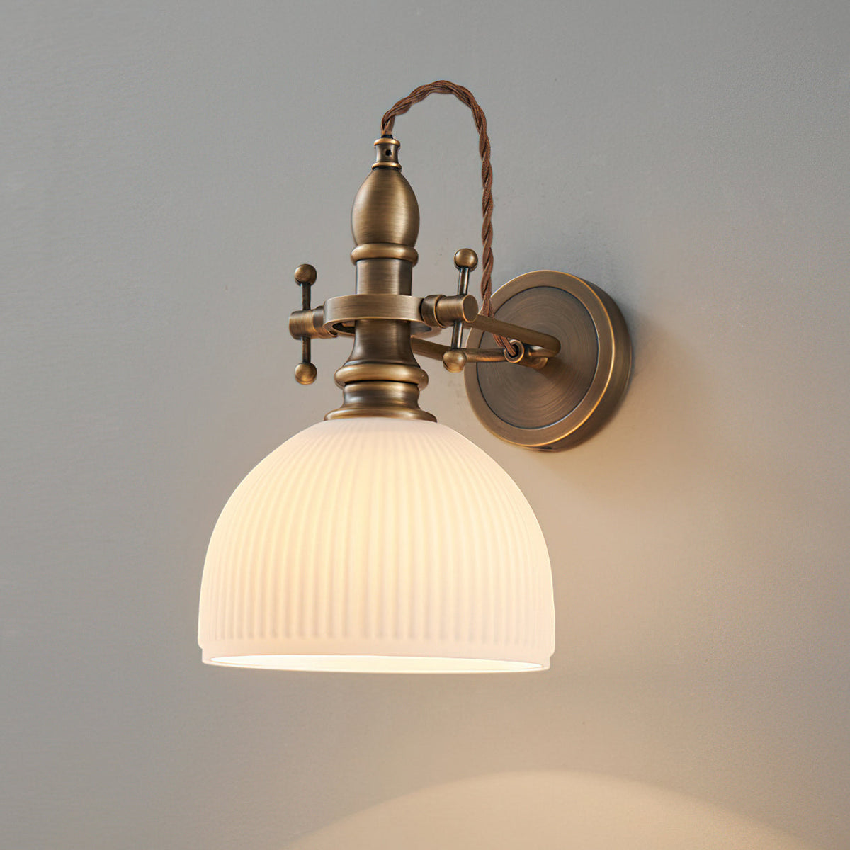 Ribbed white dome shade lit from within on a Clérachic wall light, its twisted brown cord arching over the brass stem.