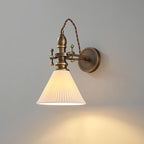 Pleated white cone shade lit on a Clérachic antique brass wall light, with the light falling down the grey wall.