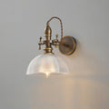 Textured clear-glass dome shade glowing around a bulb on a Clérachic brass wall light, seen straight on against grey.