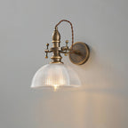 Textured clear-glass dome shade glowing around a bulb on a Clérachic brass wall light, seen straight on against grey.