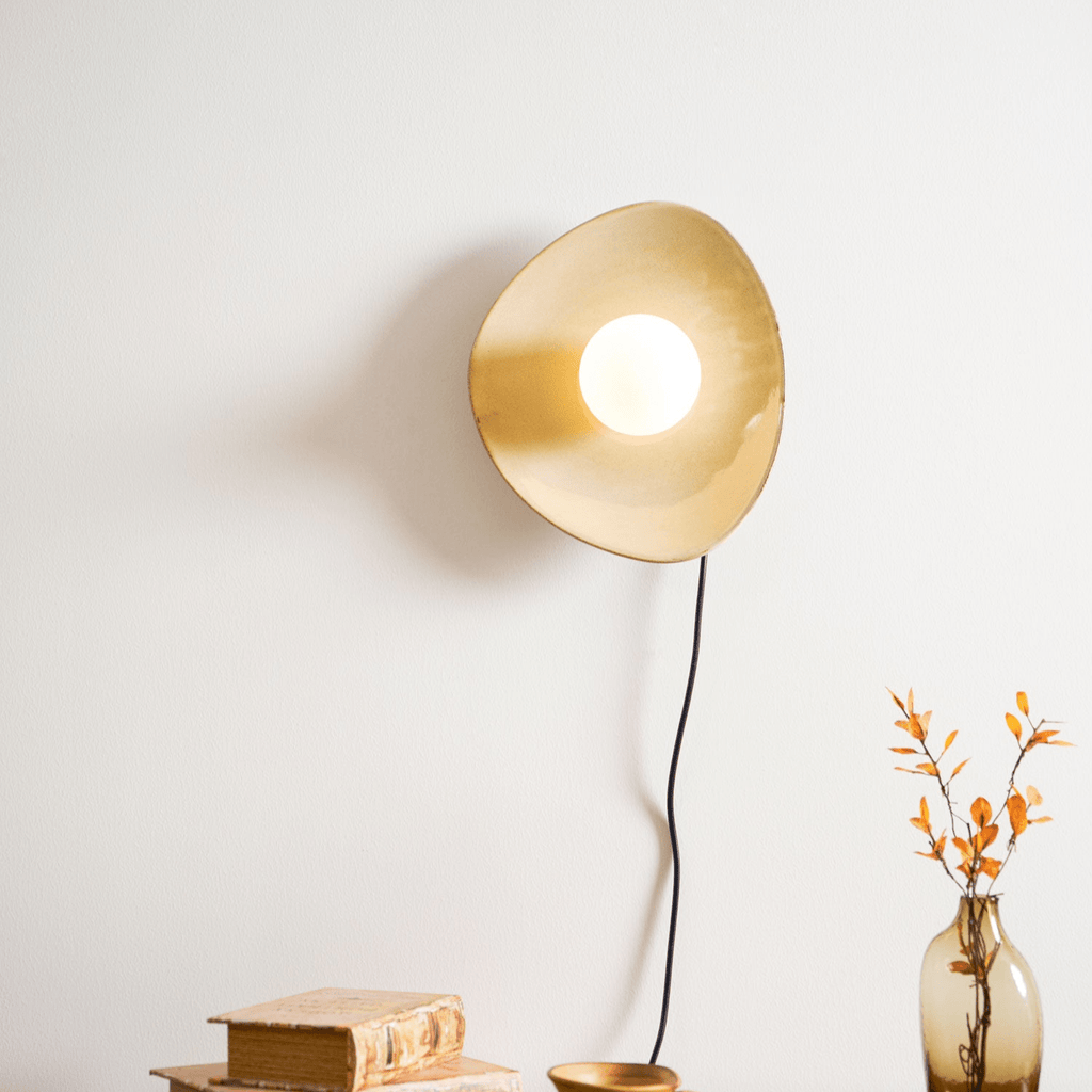 The globe lit, this Clérachic ceramic wall light glows warm gold on a white wall, with a black cord, old books and a vase of orange leaves below.