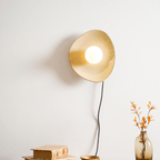 The globe lit, this Clérachic ceramic wall light glows warm gold on a white wall, with a black cord, old books and a vase of orange leaves below.