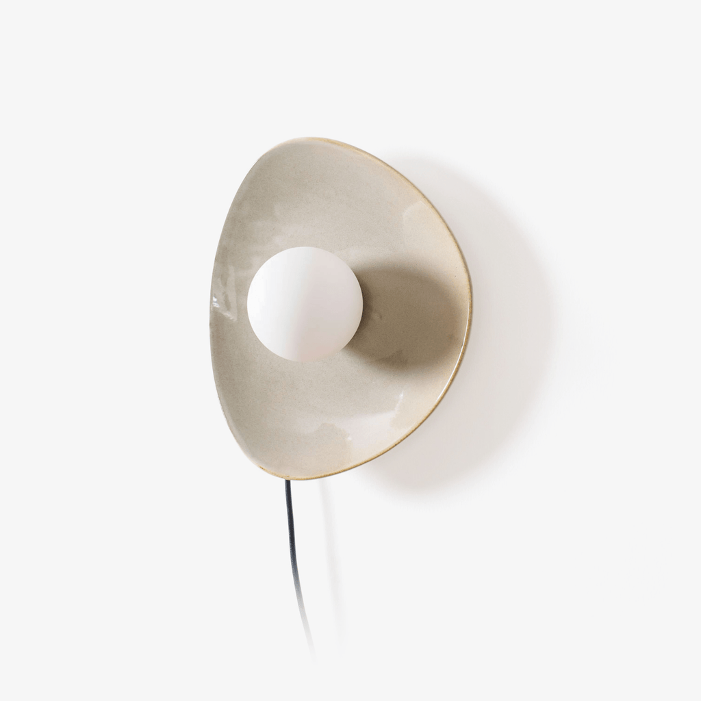 A Clérachic plug-in wall light with a taupe ceramic disc, a white globe bulb and a black cord trailing down, on a plain white backdrop.