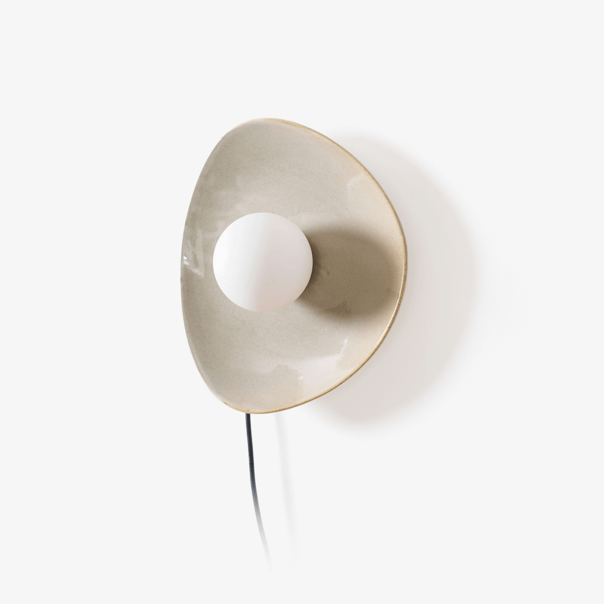 A Clérachic plug-in wall light with a taupe ceramic disc, a white globe bulb and a black cord trailing down, on a plain white backdrop.