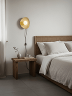A Clérachic ceramic wall light lit beside a rattan headboard, with a black cord running down to a rough wooden bedside table.