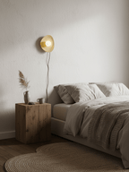 A small lit Clérachic wall light hangs on a textured plaster wall above a bed with a knitted throw and a block-style wooden table.