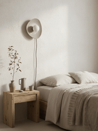 Unlit, a taupe ceramic wall light from Clérachic hangs above a bedside table with dried stems and a stoneware jug, a cord trailing down.