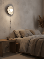 Lit warm in a quiet bedroom, this Clérachic ceramic wall light casts a glow over a textured wall, a bed and a dark wood table.