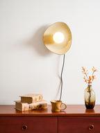 A Clérachic ceramic wall light lit warm gold above a red-brown chest of drawers with a ceramic mug, old books and a vase of orange leaves.