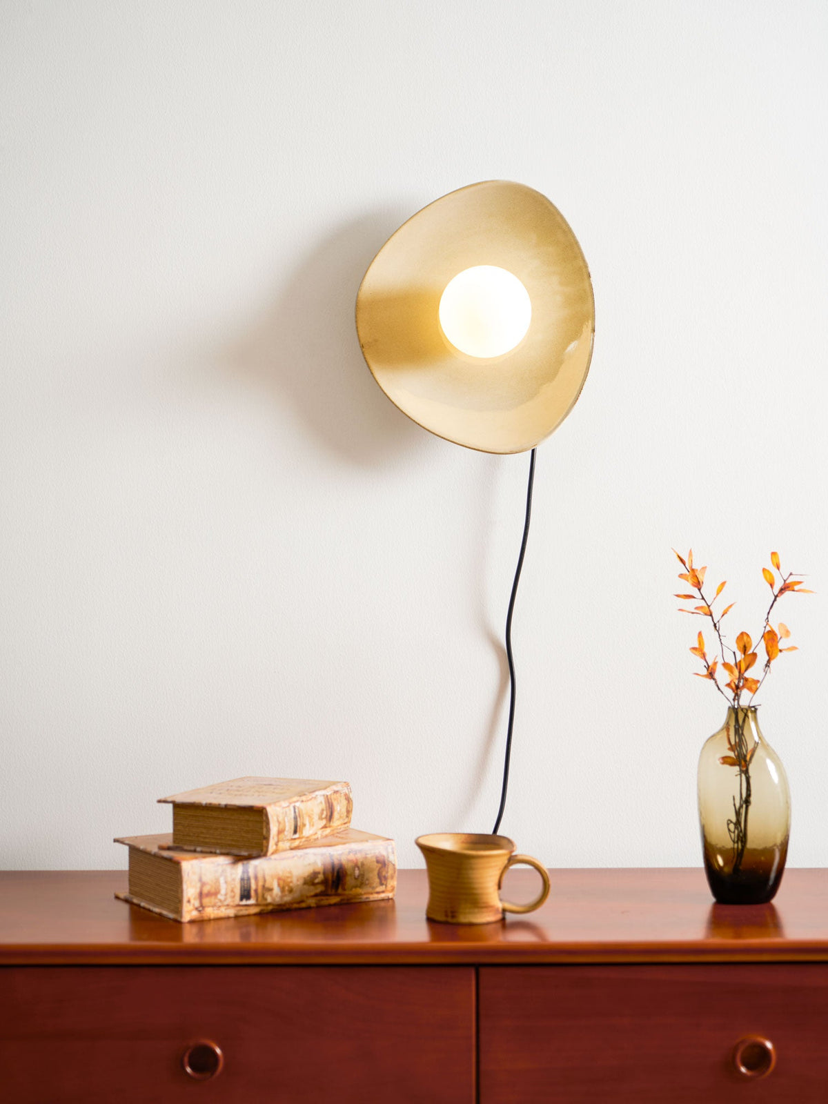 A Clérachic ceramic wall light lit warm gold above a red-brown chest of drawers with a ceramic mug, old books and a vase of orange leaves.
